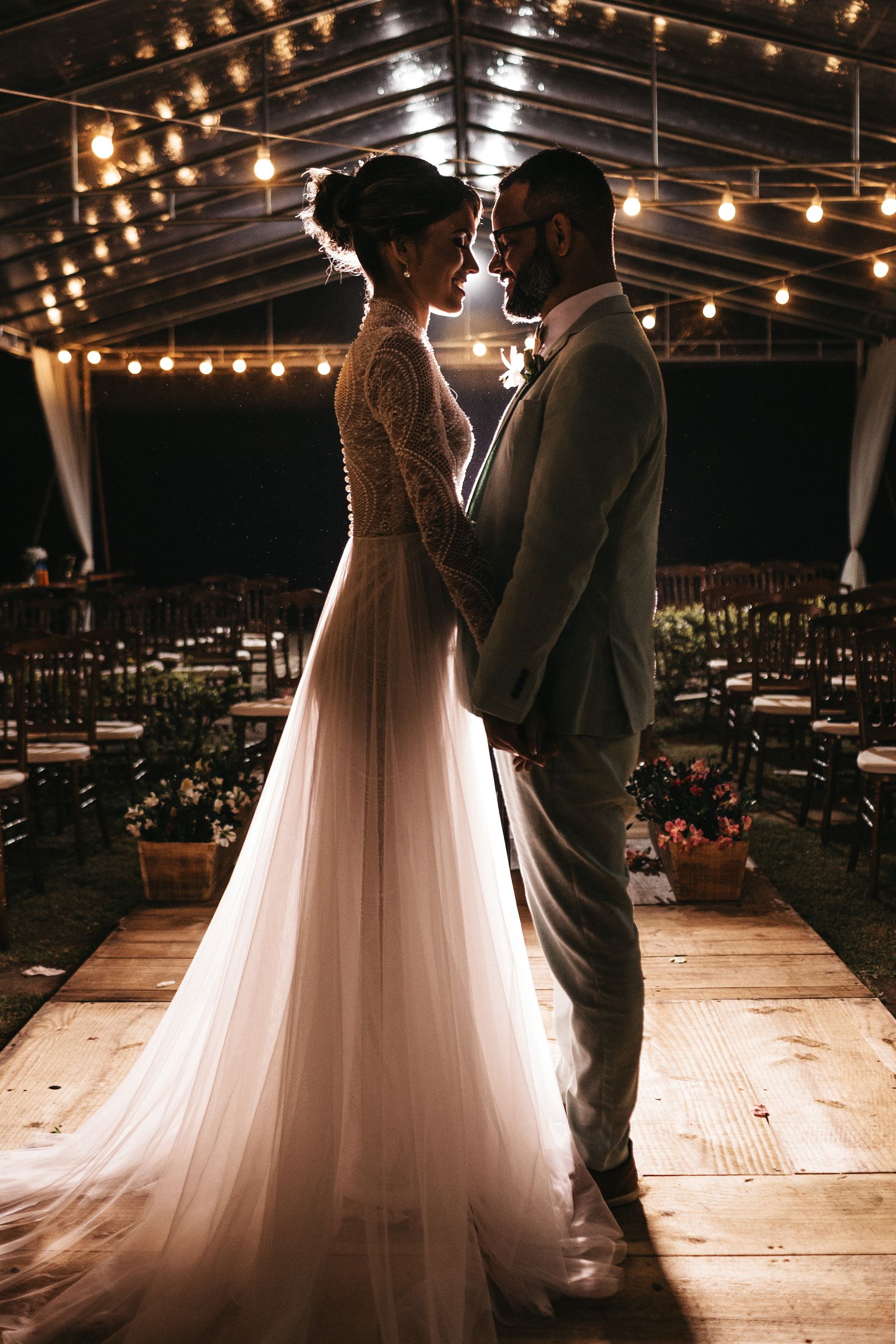 A bride in an updo and a long tulle gown, standing with the groom under string lights.