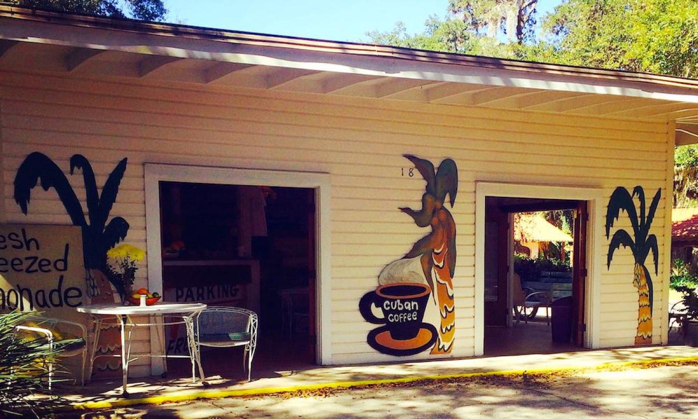 The Fountain of Juice walk-up windows on cream clapboard, with a Cuban coffee mural and a table of citrus under the oaks.