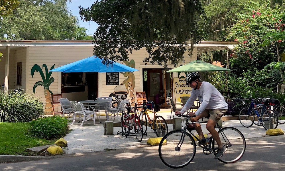 Bicycling past Fountain of Juice under live oaks and Spanish moss, with blue and green umbrellas at the outdoor tables.