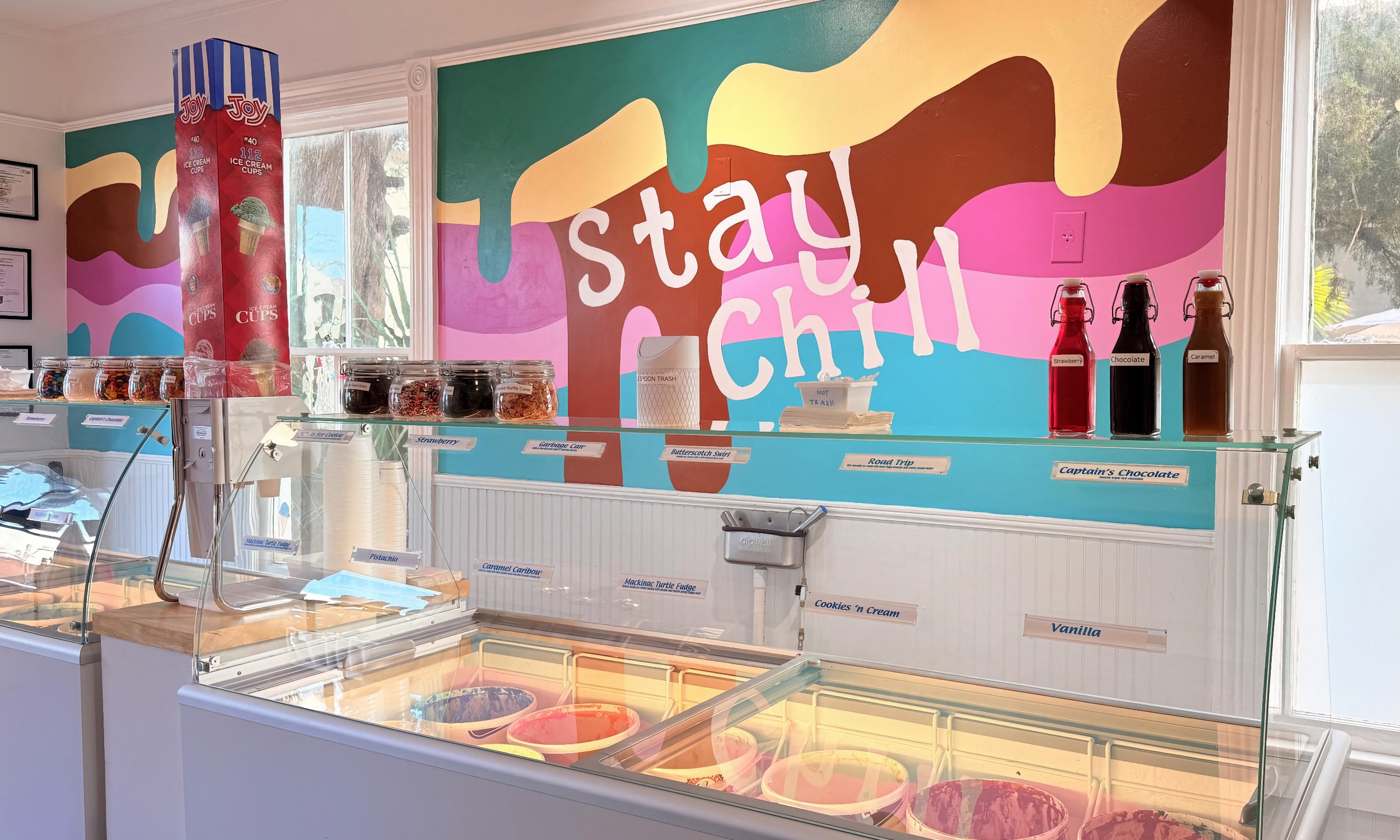 Our ice cream case under the Stay Chill mural, with topping jars and strawberry, chocolate, and caramel bottles