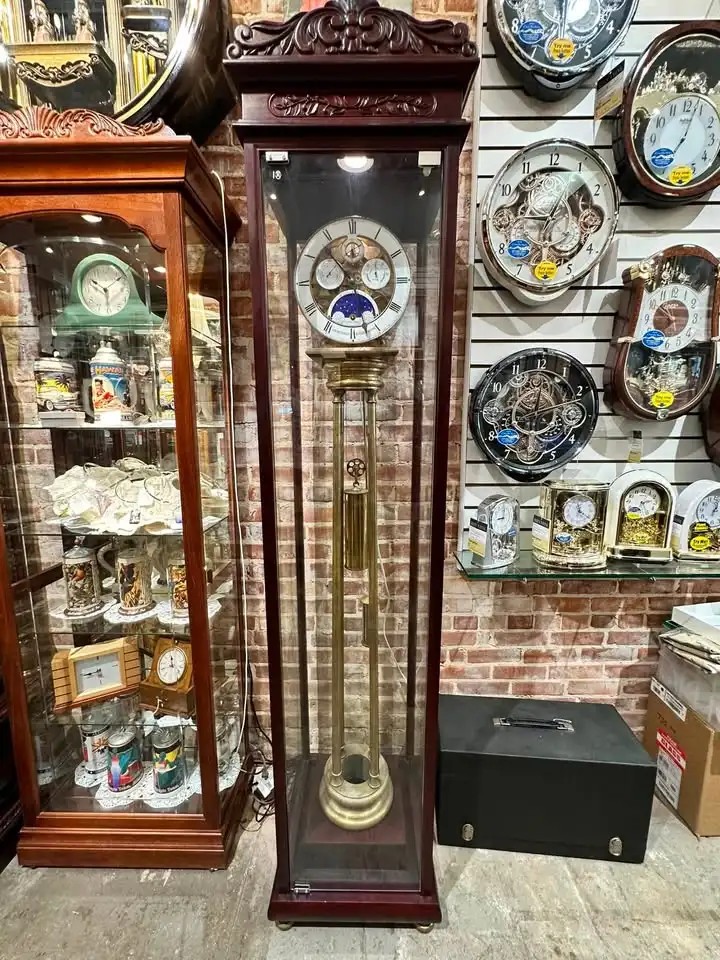 Mahogany tall-case clock with a moon-phase dial beside Rhythm wall clocks.