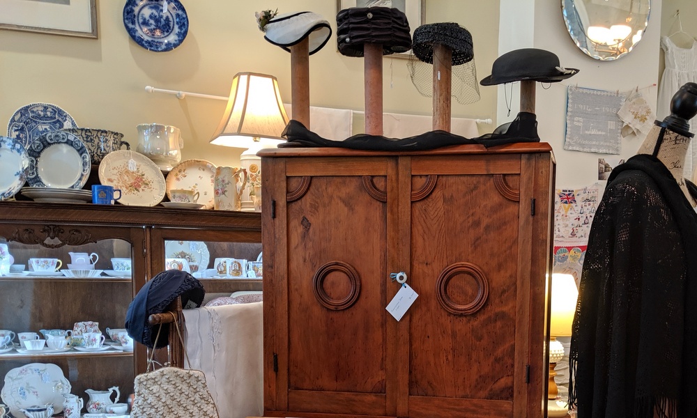 Hats on stands above a wooden cabinet, with blue and white china and teacups to the left.